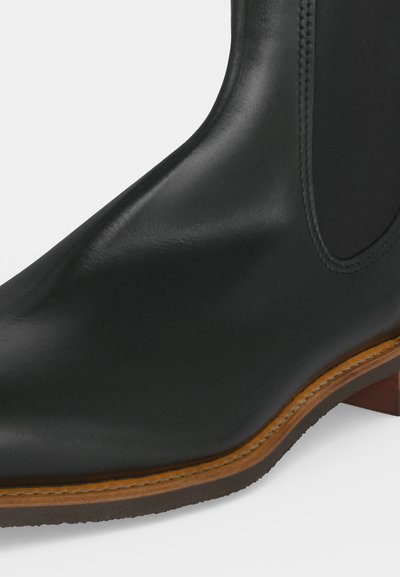 Bottine noire en cuir lisse avec semelle marron cousue et petit talon en bois, montrant un détail latéral et un panneau élastique.