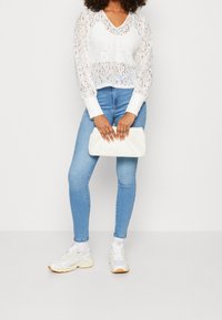 Top bianco in pizzo a maniche lunghe con scollatura a V, abbinato a jeans skinny azzurri chiari e sneakers bianche, mentre tiene una pochette bianca.