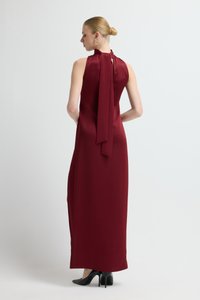 Bordowa sukienka maxi z satyny bez rękawów, z wysokim kołnierzem, ozdobiona kokardą z tyłu oraz rozcięciami po bokach. Całość dopełniają czarne szpilki.