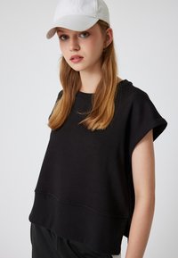 Zwarte top met korte mouwen en een losse pasvorm, gemaakt van zachte stof. Heeft een geribbelde halslijn en onderzoom. Het model draagt een witte pet.
