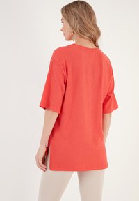 Oversized korálové tričko s krátkými rukávy, texturovaná látka, boční rozparky a kulatý výstřih. Nosené s světlými kalhotami.