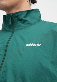 Muž v zelenej bunde na zips s bielym vyšívaným logom Adidas na hrudi na jednoduchom pozadí.