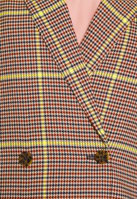 Gros plan d'un blazer à carreaux avec un motif pied-de-poule bleu marine, marron, jaune et blanc, des lignes jaunes en carreaux et des boutons en écaille de tortue.
