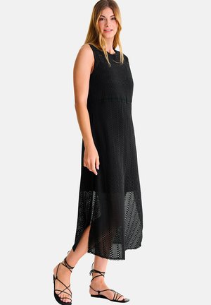 Vrouw die een mouwloze zwarte gehaakte midi-jurk draagt en zwarte sandalen met bandjes en platte zool, staand tegen een effen lichte achtergrond.