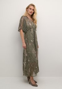 Robe longue et fluide de couleur verte avec des motifs de feuilles abstraits de teintes claires. Tissu transparent, manches courtes évasées et décolleté en V. Associée à des talons beiges.