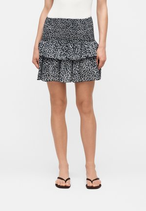 Femme portant une mini-jupe à volants noire et blanche à motif floral, un haut blanc et des tongs noires, vue de la taille aux pieds.