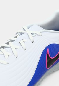 Αθλητικά παπούτσια Nike με λευκό συνθετικό πάνω μέρος, μπλε και ροζ λεπτομέρειες, και υφή στο πλάι. Λευκές κορδόνες και γκρι σόλα.