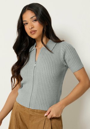 Femme aux longs cheveux foncés portant un pull gris côtelé à manches courtes avec fermeture éclair et un pantalon marron sur un fond uni.