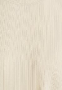Žebrovaný top v jasně krémové barvě, s vertikálními pruhy, měkkou texturou a kulatým výstřihem. Materiál vypadá lehce a pružně.