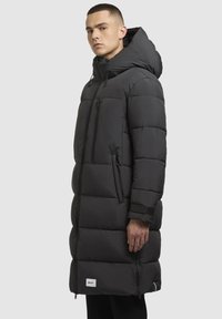 Lang sort pufferjakke med hætte, der har lynlås foran, sidelommer og elastiske manchetter. Materialet er glat og polstret for isolering.