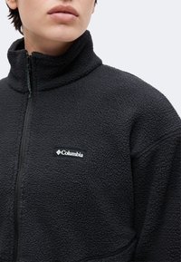 Άτομο που φοράει μαύρο, υφασμάτινο fleece μπουφάν με ψηλό γιακά και ορατό λογότυπο Columbia στο στήθος.