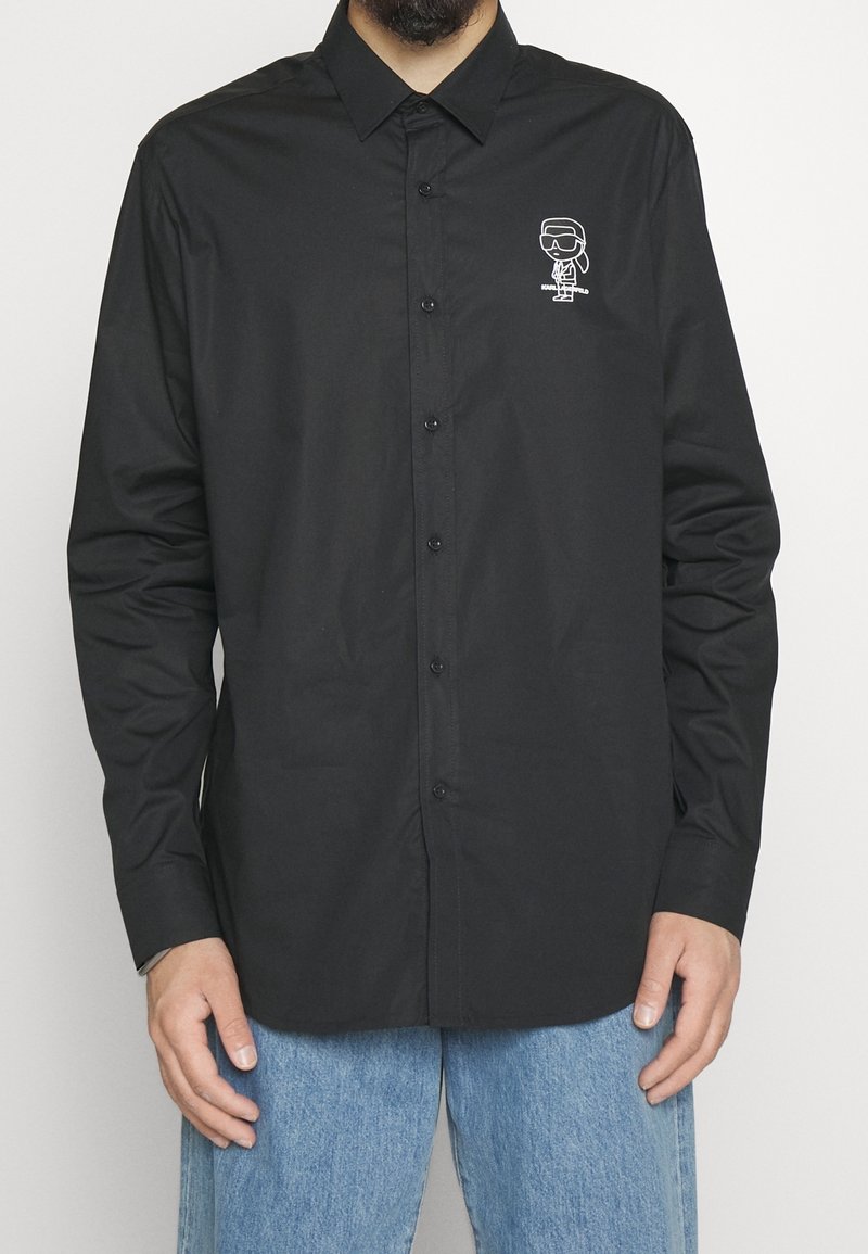 Homme portant une chemise noire à boutons avec un petit logo cartoon blanc sur la poitrine et un jean bleu clair sur fond uni.