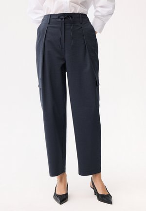 Marineblaue, hoch taillierte Hose mit Tunnelzugbund, seitlichen Cargotaschen, kombiniert mit schwarzen Slingback-Pumps mit spitz zulaufender Schuhspitze und weißer Bluse.