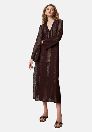 Donna in piedi che indossa un lungo vestito marrone scuro all'uncinetto con maniche lunghe e sandali piatti con punta aperta.