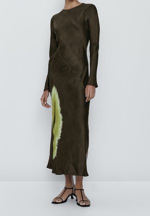 Kobieta w długiej sukience midi o ciemnooliwkowym kolorze z dużym, jasnym zielonym i żółtym kwiatowym nadrukiem po boku, w czarnych sandałach z paskami.