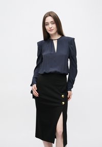 Granatowa bluzka z dekoltem w kształcie łezki i marszczonymi ramionami, zestawiona z czarną spódnicą z bocznym rozcięciem i trzema złotymi guzikami jako ozdobą.