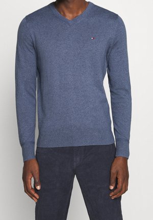 Uomo che indossa un maglione blu a scollo a V con un piccolo logo a bandiera rossa, bianca e nera sul petto e pantaloni scuri, in piedi davanti a uno sfondo semplice.