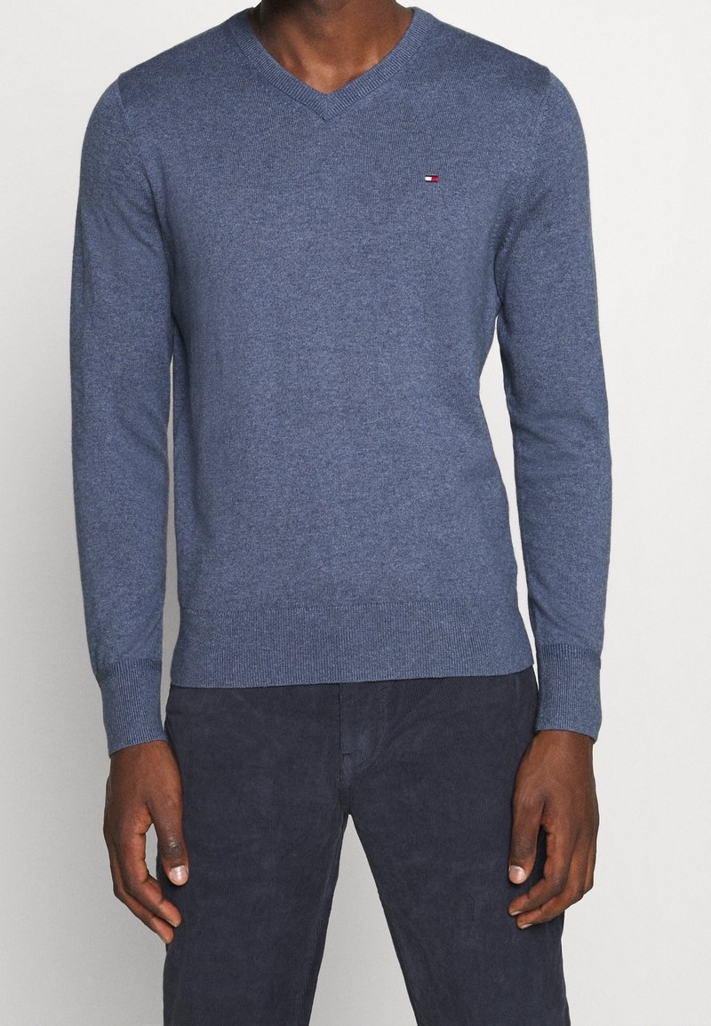 Homme portant un pull bleu à col en V avec un petit logo de drapeau rouge, blanc et noir sur la poitrine et un pantalon foncé, debout devant un fond uni.