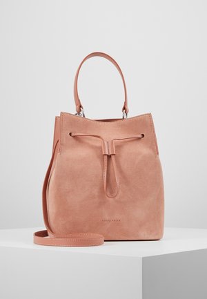 Weiche rosa Wildleder-Handtasche mit obenliegendem Griff, verstellbarem Schulterriemen und Kordelzugverschluss auf einem weißen Podest.