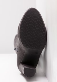 Bottine en daim gris foncé posée la semelle vers le haut contre des murs blancs, montrant la semelle en caoutchouc texturée et le talon bloc empilé.