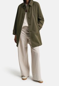 Osoba v olivově zeleném kabátu na knoflíky, bílém svetr s pleteninou, béžových kalhotách širokého střihu a bílých teniskách se hnědými podrážkami.