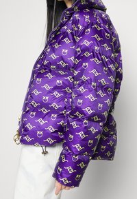 Paarse pufferjas met een glanzende afwerking, voorzien van een herhalend geel logopatroon, ritssluiting en trekkoorden aan de zoom en manchetten.