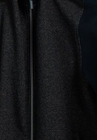 Getextureerde donkere jas met een ritsluiting; beschikt over een geweven patroon van bruin en zwart. Inclusief contrasterende accenten van gladde stof.