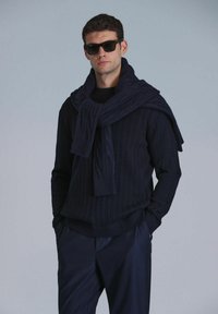Marineblaue Kabelstrickpullover kombiniert mit dunklen Hosen und einem übergroßen Schal, der über die Schultern drapiert ist; das Model trägt dunkle Sonnenbrillen.