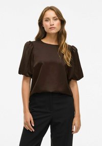 Vrouw met lang golvend haar, gekleed in een donkerbruine blouse met pofmouwen en zwarte broek, staande voor een effen witte achtergrond.