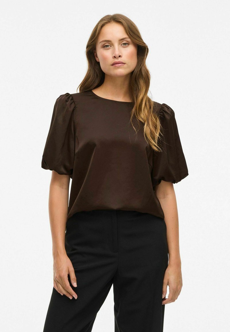 Vrouw met lang golvend haar, gekleed in een donkerbruine blouse met pofmouwen en zwarte broek, staande voor een effen witte achtergrond.