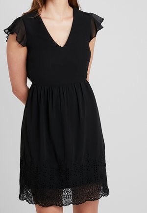 Femme portant une robe noire avec des manches volantes transparentes, un décolleté en V, une taille froncée et un ourlet en dentelle florale découpée.