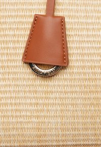 Tan leather accent with stitching attached to a circular metal ring, set against a textured beige woven material.