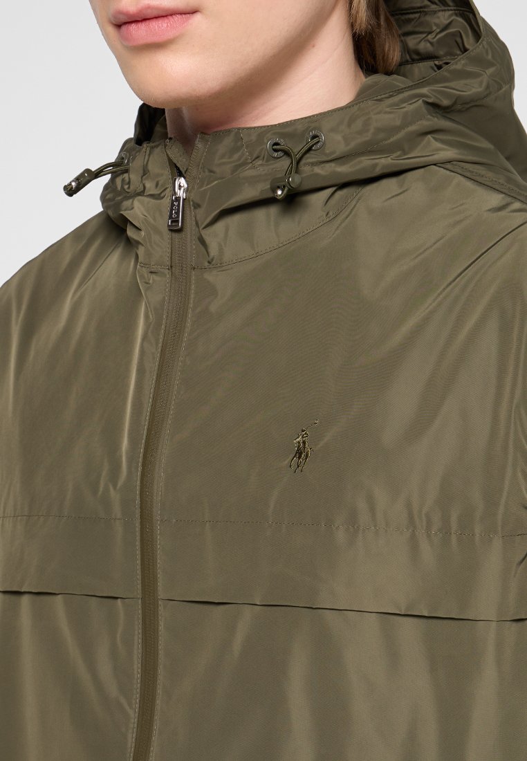 Olivegrüne wasserdichte Jacke mit Reißverschluss, verstellbarem Zugkordelkapuze und einem Logo auf der Brust. Glattes, leichtes Material.
