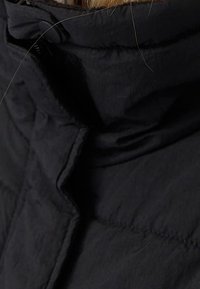 Zwarte pufferjack met hoge kraag, gemaakt van een gladde, textuurstof met stikdetails langs de naden. Veilige knoopsluiting bij de nek.