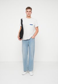 Tricou gri din bumbac cu un logo albastru pe piept, asortat cu blugi drepți de culoare albastră deschisă și adidași albi; modelul ține o geantă neagră.
