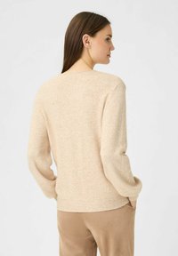 Beiger Strickpullover mit rundem Halsausschnitt, langen Ärmeln und geripptem Saum. Weiche Textur und minimalistisches Design. Kombiniert mit beigen Hosen.
