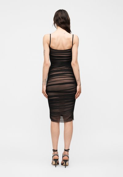 Γυναίκα που στέκεται με την πλάτη γυρισμένη, φορώντας μια εφαρμοστή μαύρη midi φόρεμα με πιέτες και λεπτές τιράντες, μαζί με μαύρα ψηλοτάκουνα σανδάλια.