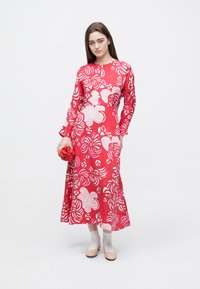 Rotes Maxikleid mit Blumenmuster, langen Ärmeln und ausgestelltem Saum. Glatter Stoff, kombiniert mit einer roten Clutch, unterstreicht das lebendige Design.