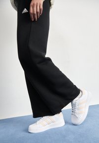 Vita Adidas-sneakers med rosa detaljer, som har tre ränder på sidan och en texturerad gummisula, ihop med svarta mjukisbyxor.