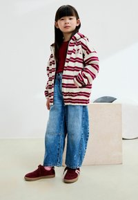 Veste à capuche rayée en crème et bordeaux, associée à un jean en denim bleu ample et des baskets bordeaux. Présente une coupe décontractée et des poches spacieuses.
