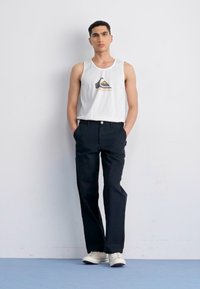 Witte katoenen tanktop met logo, gecombineerd met zwarte op maat gemaakte broeken en witte sneakers. Model staat met handen in de zakken op een blauwe vloer.