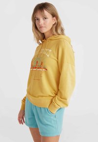 Felpa gialla con una stampa grafica che presenta figure e una tavola da surf. Abbinata a pantaloni corti azzurri. Tessuto morbido, vestibilità casual.