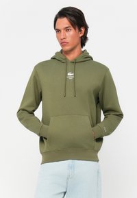 Olijfgroene hoodie met een kangoeroezak, voorzien van een wit Lacoste-logo op de borst en ribgebreide mouwen, gemaakt van zachte stof.