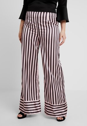 Vrouw draagt een broek met hoge taille en wijde pijpen met verticale roze en zwarte strepen, gecombineerd met een zwarte top en zwarte open-toe schoenen.