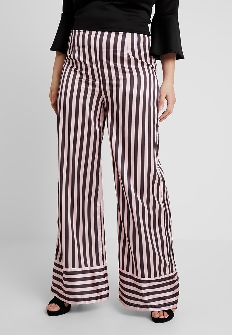Femme portant un pantalon taille haute à jambes larges avec des rayures verticales rose et noir, associé à un haut noir et des chaussures noires à bouts ouverts.