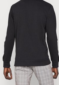 Felpa nera con maniche lunghe, polsini e orlo a coste, tessuto liscio, mostrata da dietro, indossata con pantaloni a quadri.
