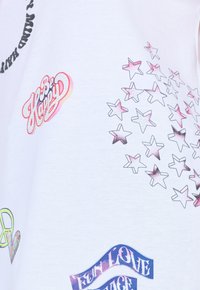 T-shirt blanc avec des impressions graphiques colorées incluant les phrases "Soyez Heureux" et "AMOUR AMUSANT", ainsi que des formes d'étoiles de tailles et nuances variées.