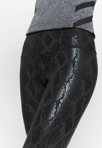 Nära bild av svarta leggings med glänsande ormskinnsmönster, iförda med en grå, kort, figurnära topp mot vit bakgrund.