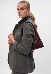 Burgundska kožna torba s jedinstvenim trokutastim oblikom, glatkom teksturom i vidljivim šavovima, nošena uz kaput s uzorkom riblje kosti.