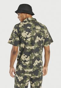 Camicia a maniche corte con motivo camouflage nei toni del verde e del nero, con colletto e tessuto testurizzato. Cappello a secchiello nero.